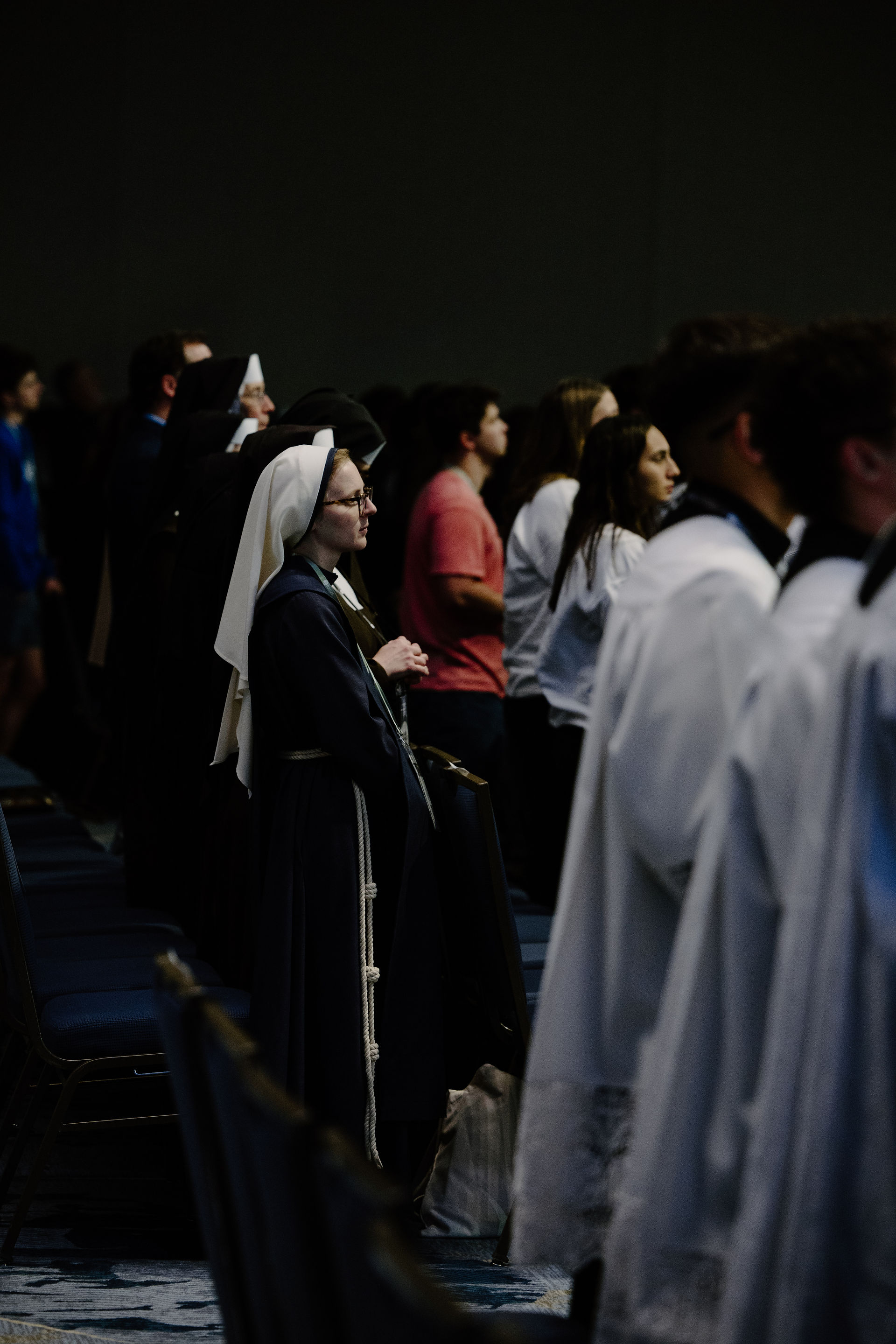 Nuns and people stand in prayer in a dimly lit room. Nuns wear traditional habits, and attendees are dressed casually. Quiet, reverent mood.