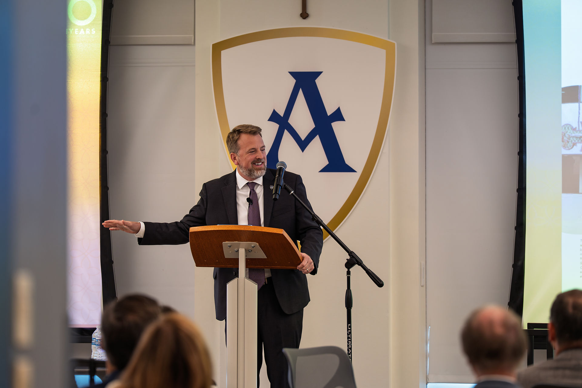 Man in a suit speaking at a podium with a microphone, shield logo on wall behind, audience in foreground, setting is a presentation room.