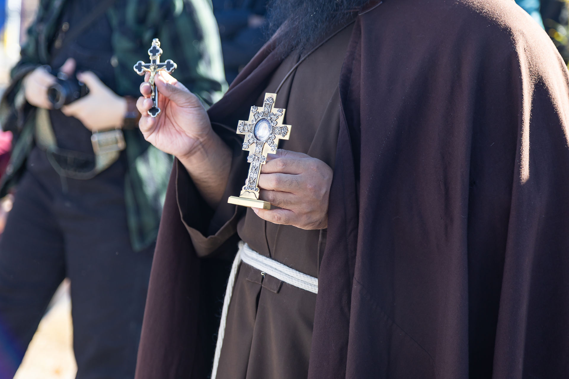 Person in brown robe holding two ornate crosses, one silver and one gold. A camera is seen in the background, with sunlight casting shadows.
