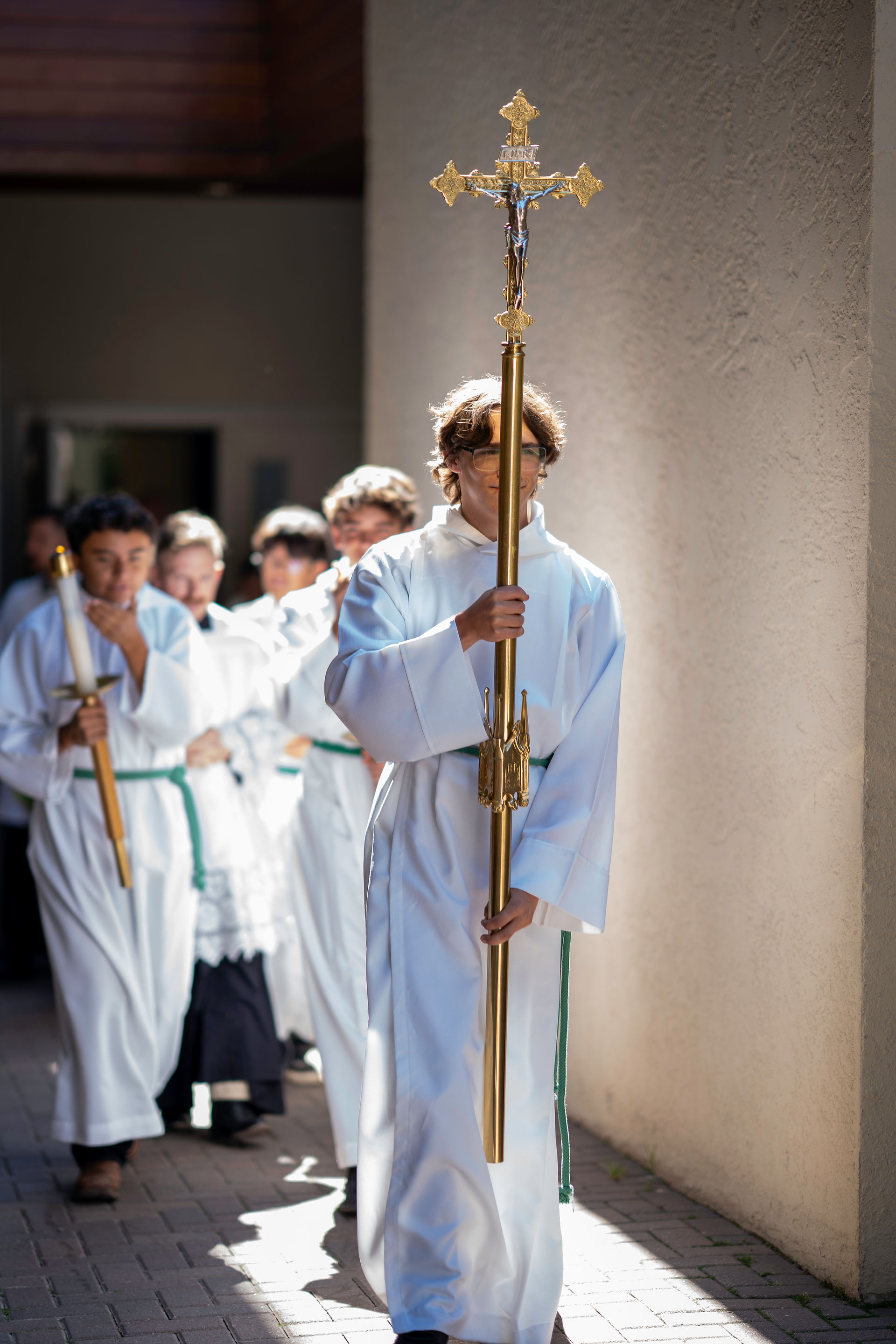 A group of altar servers in white robes processes with a large gold cross in a sunlit corridor. Solemn and focused mood.