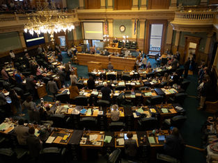 View of a legislative chamber with officials standing by desks in a grand room, large chandelier, flags, and screens. Mood is formal.