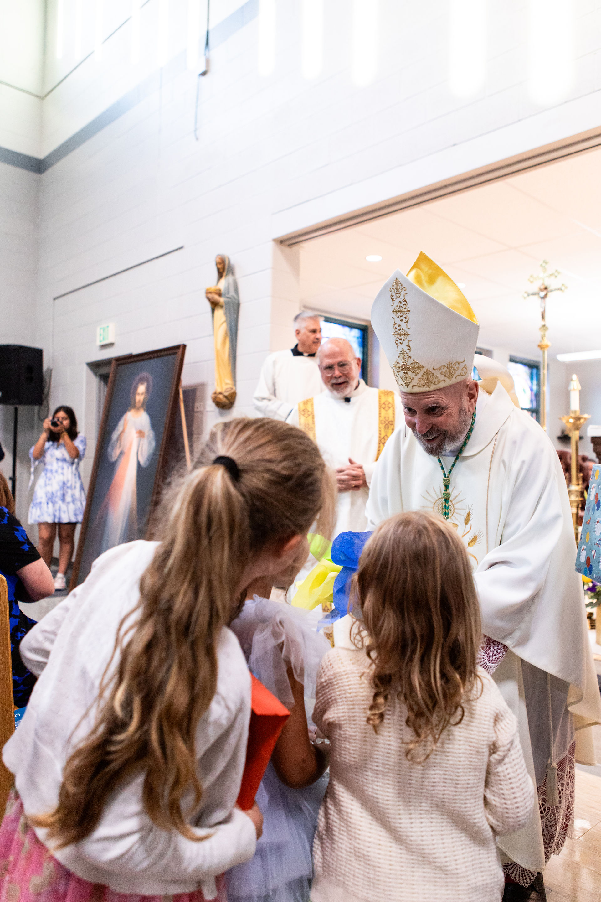 A bishop in ornate robes greets children in a church. A portrait and a statue are in the background. The mood is joyful and reverent.