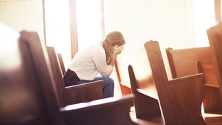A woman sits alone on a church pew, head in hands, appearing distressed. Sunlight filters through large windows, creating a warm glow.