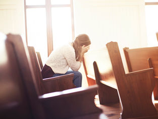 A woman sits alone on a church pew, head in hands, appearing distressed. Sunlight filters through large windows, creating a warm glow.