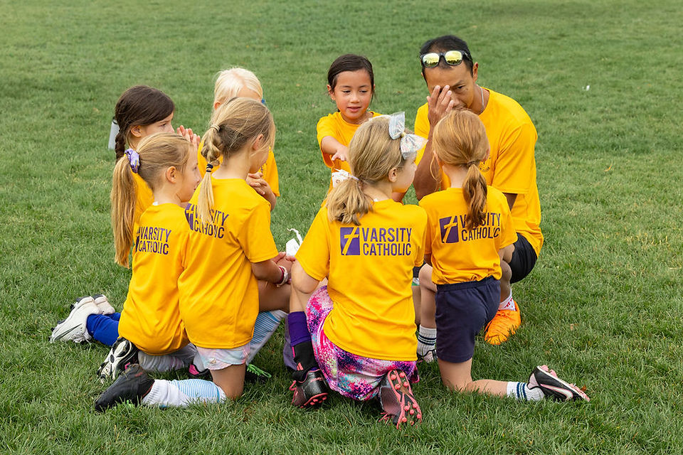 Children in yellow Varsity Catholic shirts sit in a grass huddle with a coach, listening during an outdoor team talk.