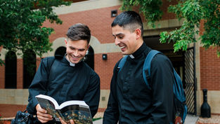 Two men in black clerical clothing smile while reading a book outdoors against a brick building and green trees. One carries a backpack.