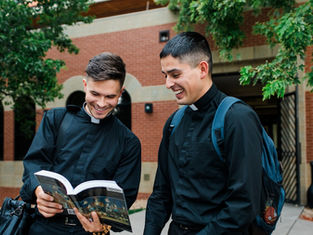 Two men in black clerical clothing smile while reading a book outdoors against a brick building and green trees. One carries a backpack.