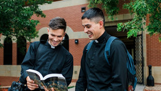 Two men in black clerical clothing smile while reading a book outdoors against a brick building and green trees. One carries a backpack.