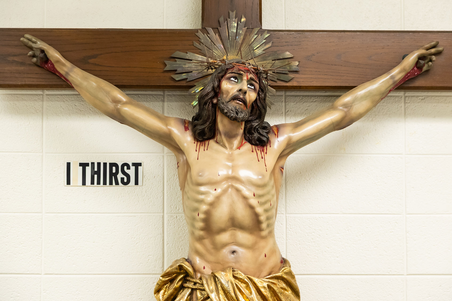 A crucifix statue of Jesus with a crown of thorns, bleeding wounds, and golden cloth. "I Thirst" text on white wall background. Somber mood.