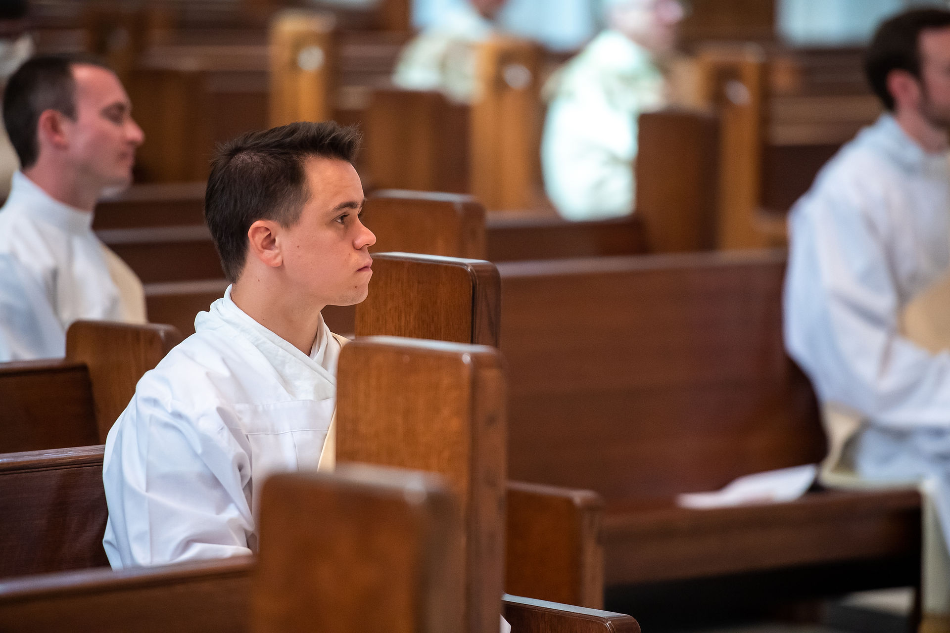 Young man in white robe sits in a wooden church pew, gazing forward attentively. Dim interior with blurred figures in background.