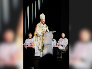 A bishop in ornate robes speaks at a clear lectern with priests seated behind him against a dark backdrop. Light streams from above.