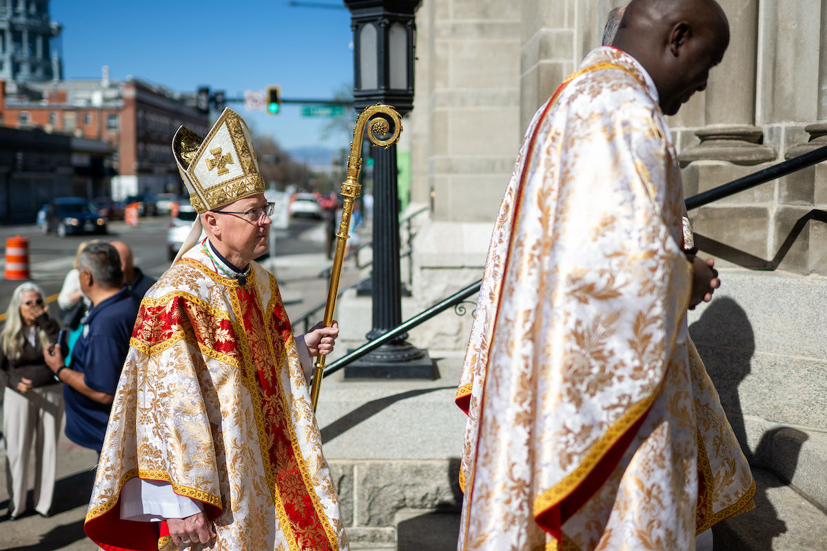 A bishop carrying a crosier walks up the steps of a cathedral alongside another priest outdoors.
