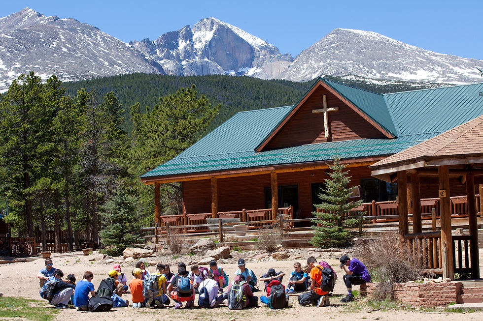 A group of people sit in a circle outside a log cabin with a cross, set against forested mountains under a clear blue sky.