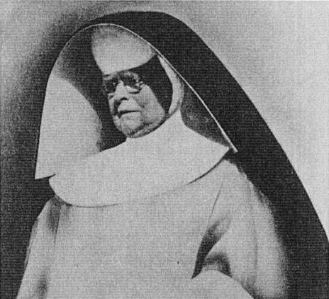 Black-and-white portrait of an elderly nun in a white habit and dark veil, looking solemn against a plain background.