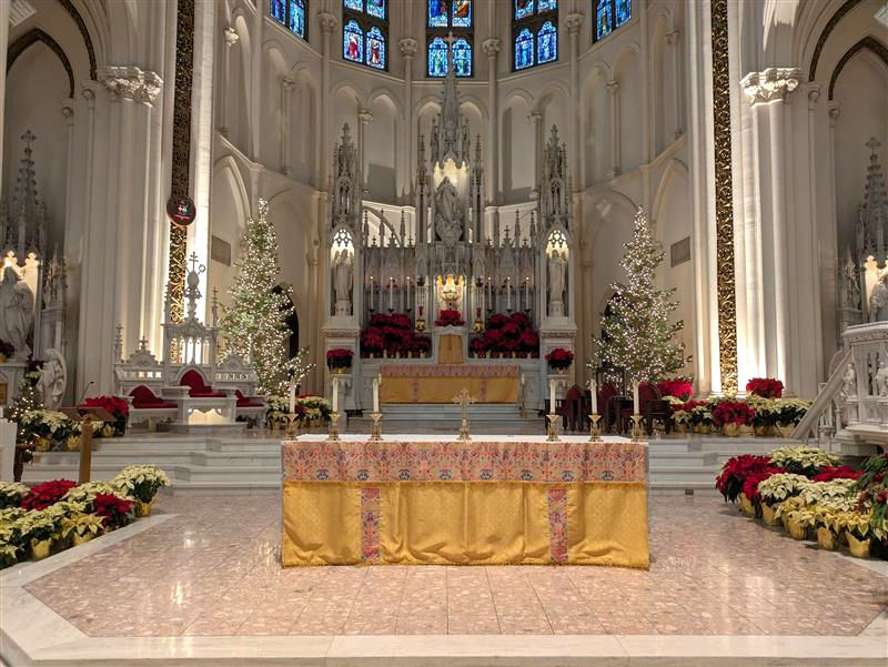Ornate church altar with a gold-cloth table, flanked by lit Christmas trees, surrounded by red and white flowers. Stained glass above.