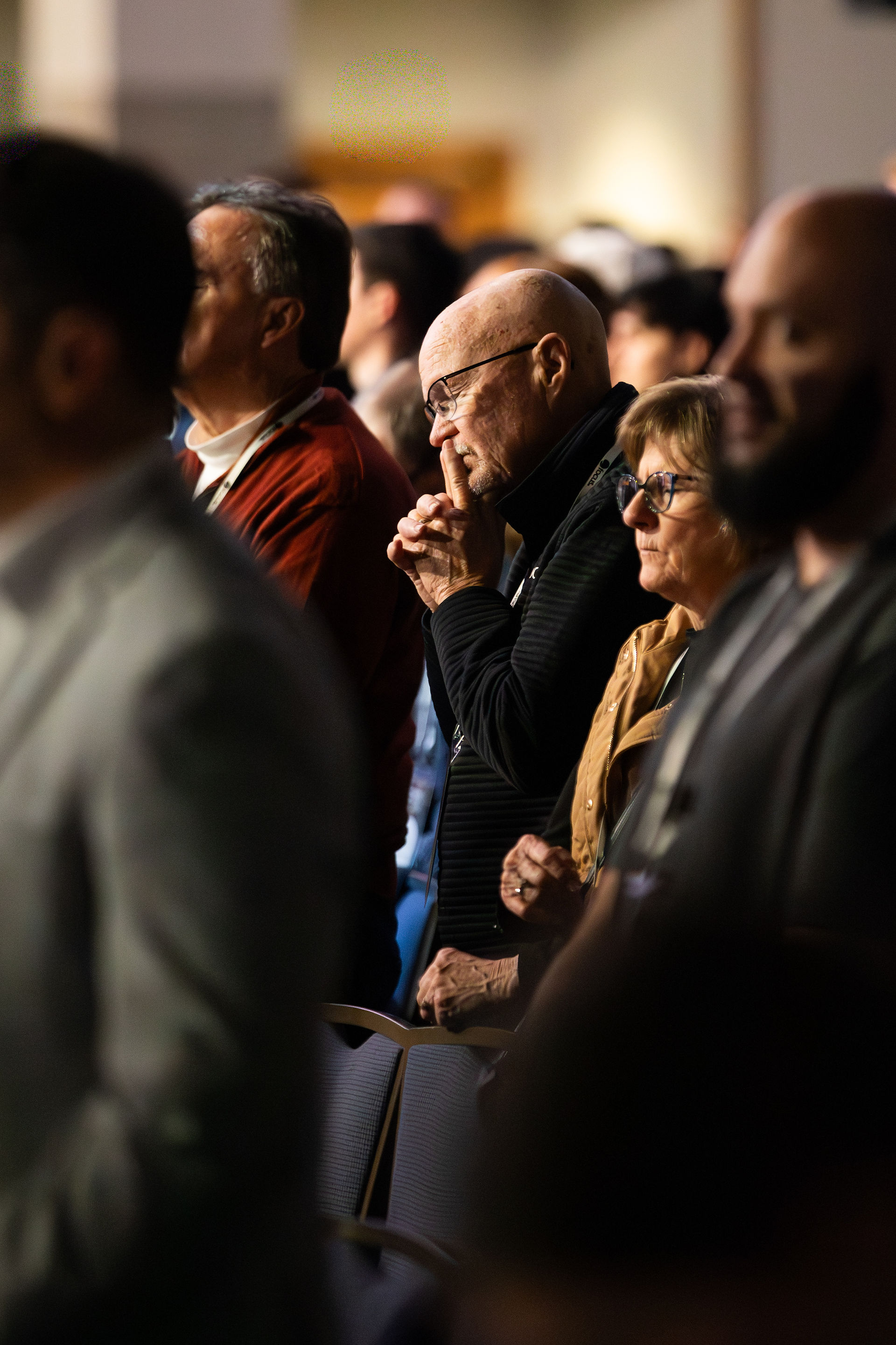 Elderly people observing attentively in a crowded indoor setting, some with hands clasped. The mood is thoughtful; lighting is warm and soft.