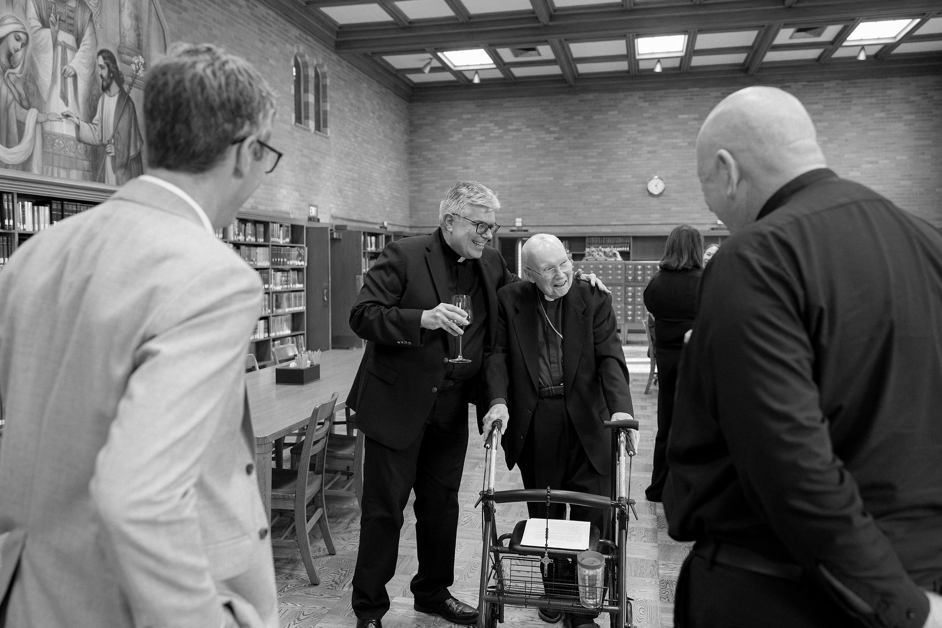 Clergymen in black attire converse in a library with large mural, bookcases, and tables. One holds a wine glass. They appear happy and relaxed.