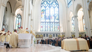 Catholic mass in a bright cathedral, priests at the altar and congregation seated beneath tall stained-glass windows.