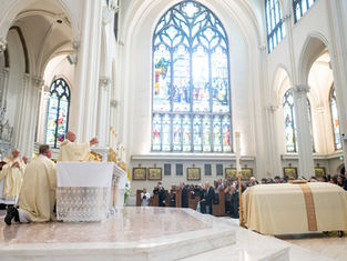 Catholic mass in a bright cathedral, priests at the altar and congregation seated beneath tall stained-glass windows.