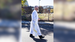 A person in a white robe walks on a sunny street, holding a rosary. Background includes a fence, trees, and distant buildings.