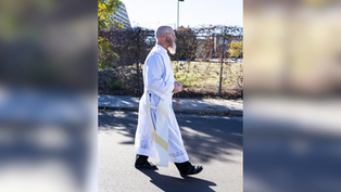 A person in a white robe walks on a sunny street, holding a rosary. Background includes a fence, trees, and distant buildings.
