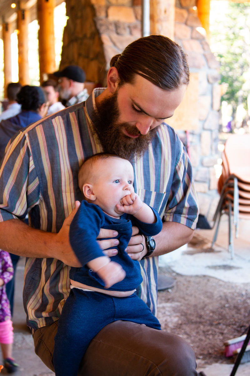 Man holding baby in blue, indoors with stone wall and chairs. Man wears striped shirt, looking down, baby sucking thumb. Warm light.