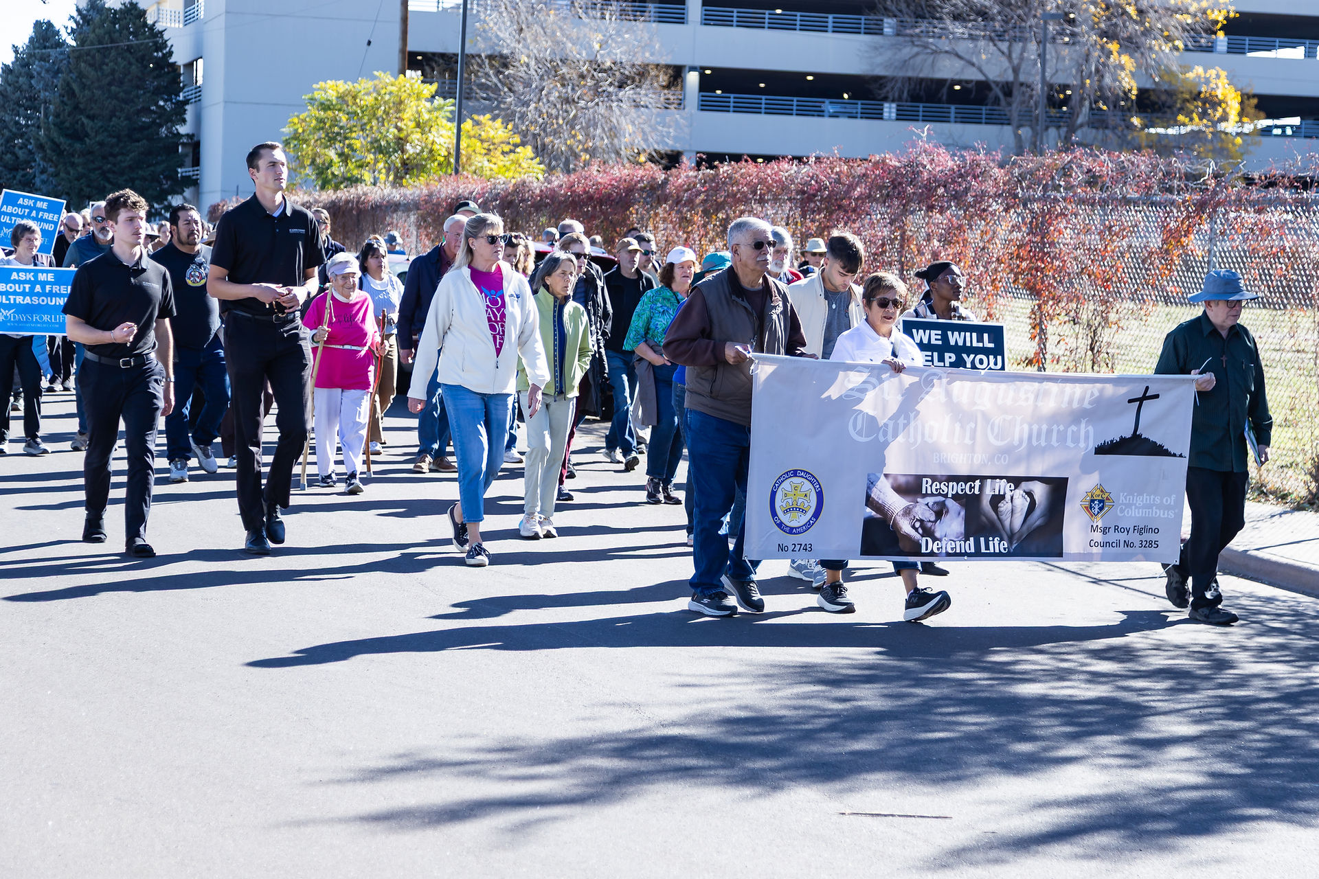 A group of people march on a sunny street holding banners with messages supporting life, set against a backdrop of autumn trees and a building.