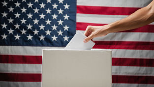 Hand placing a ballot into a box against the backdrop of a U.S. flag, symbolizing voting and patriotism.