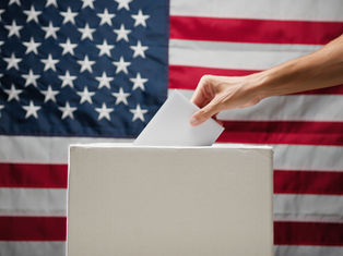 Hand placing a ballot into a box against the backdrop of a U.S. flag, symbolizing voting and patriotism.
