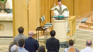 Priest in white robe speaks at a podium in a church. Congregation stands, facing altar. Cross and plants visible. Calm atmosphere.