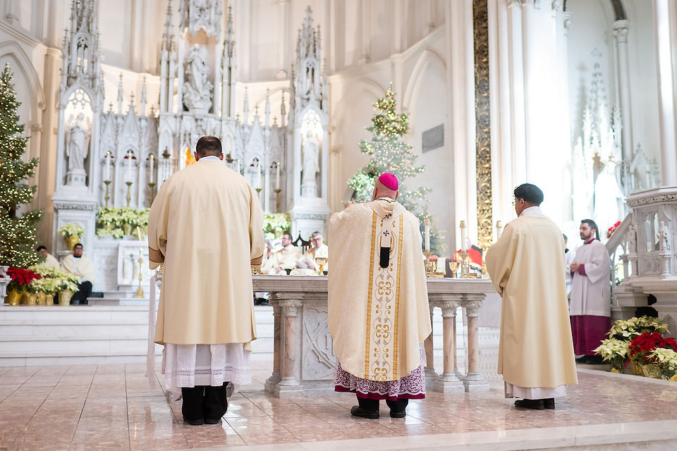 Clergy in white and gold robes conduct a ceremony in a decorated cathedral. Christmas trees, poinsettias, and an ornate altar are visible.