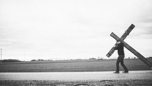 Man carrying large wooden cross along a rural road; vast field in background. Black and white image conveys a solemn, reflective mood.