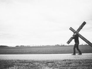 Man carrying large wooden cross along a rural road; vast field in background. Black and white image conveys a solemn, reflective mood.