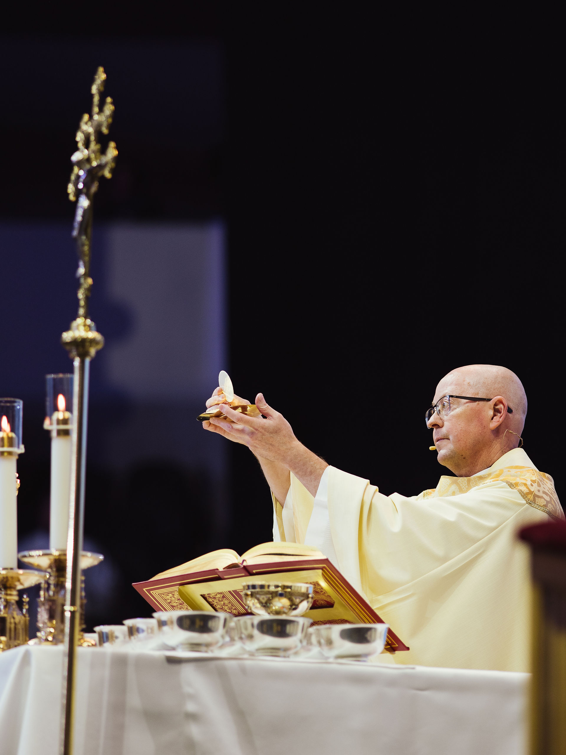 A bishop in yellow vestment raises the Eucharist near an altar with a large open book and candles, evoking a solemn, sacred mood.