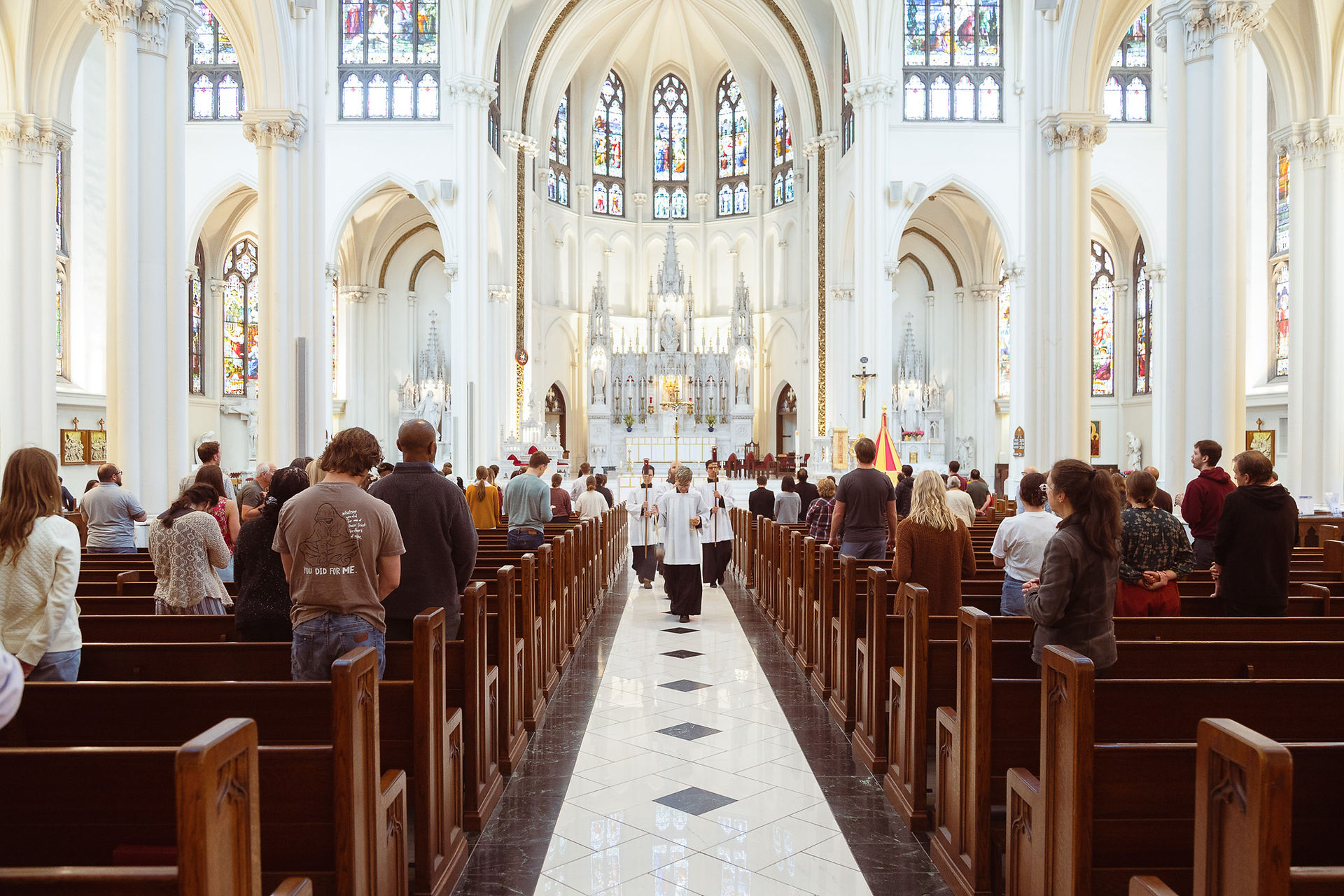 People stand in a cathedral with stained glass windows, wooden pews, and vaulted ceilings. Clergy in white robes walk down the aisle.