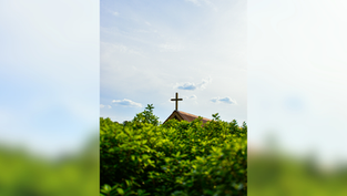 Cross on a church roof above bright green trees against a blue sky with clouds. Peaceful and serene atmosphere.