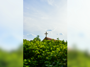 Cross on a church roof above bright green trees against a blue sky with clouds. Peaceful and serene atmosphere.