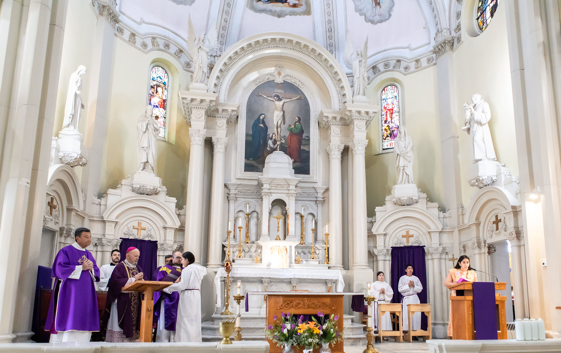 Clergy in purple robes conduct a service in a church with a decorated altar. A woman reads at a lectern. Stained glass windows in the background.