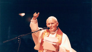 A man in religious attire speaks at a podium, holding papers. He gestures with one hand. The background is dark, creating a solemn mood.