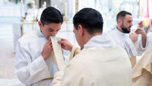 A newly ordained deacon kisses his stole as he is vested for the first time. Soft lighting and altar in the blurred background.