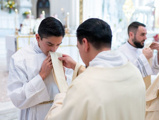 A newly ordained deacon kisses his stole as he is vested for the first time. Soft lighting and altar in the blurred background.