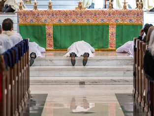 People in white robes lie prostrate before a decorated altar in a church, surrounded by seated onlookers. The atmosphere is solemn.
