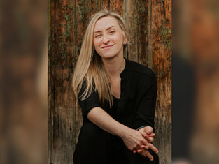 Smiling woman with long blonde hair in black outfit sits against a rustic wooden background, exuding a relaxed and cheerful mood.
