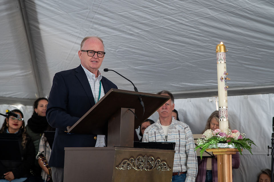 A man in a suit speaks at the podium beside a decorated Paschal candle during the church event.