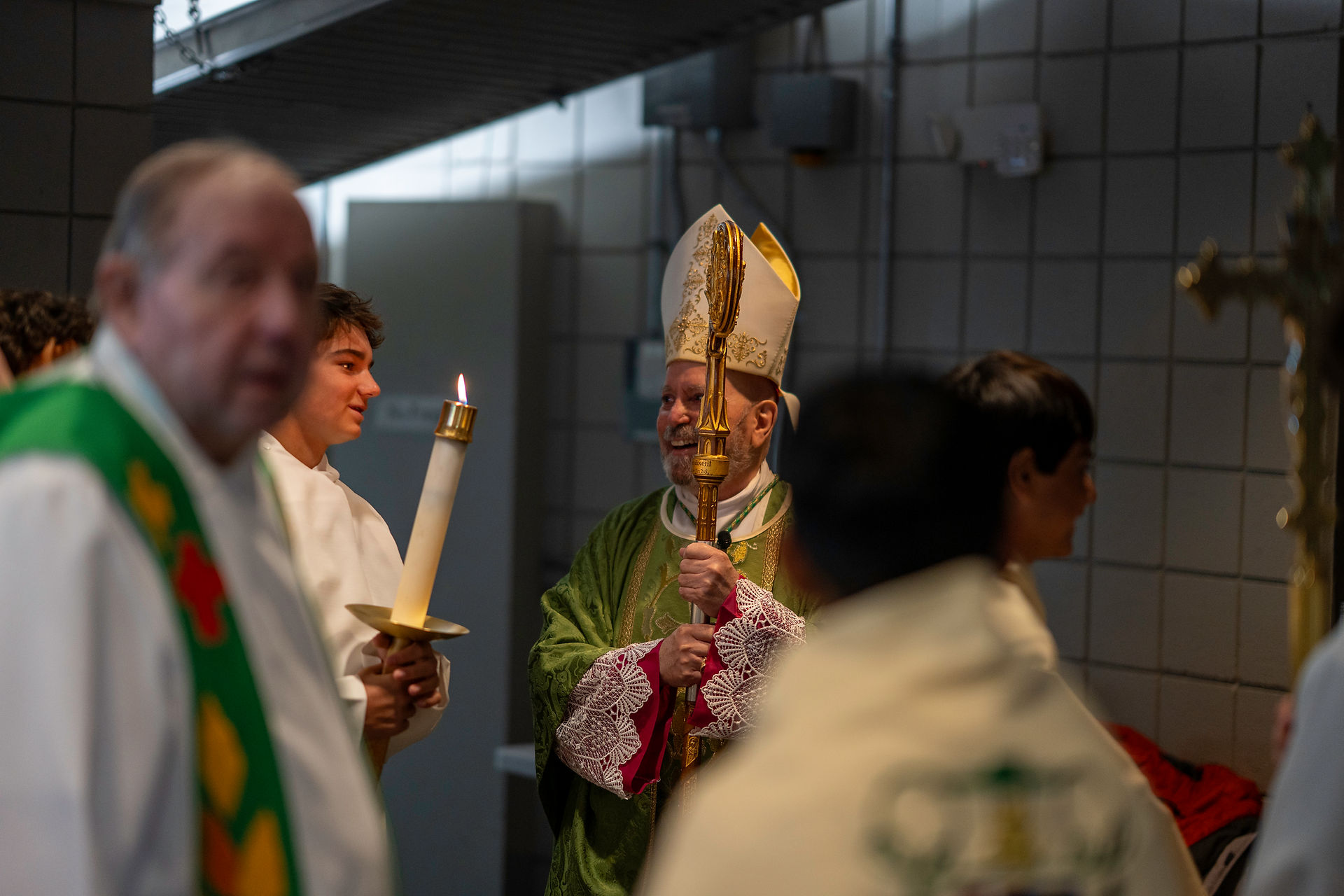 A religious ceremony featuring several individuals in liturgical vestments. One holds a candle, another wears a mitre and carries a staff, with others visible in the background.