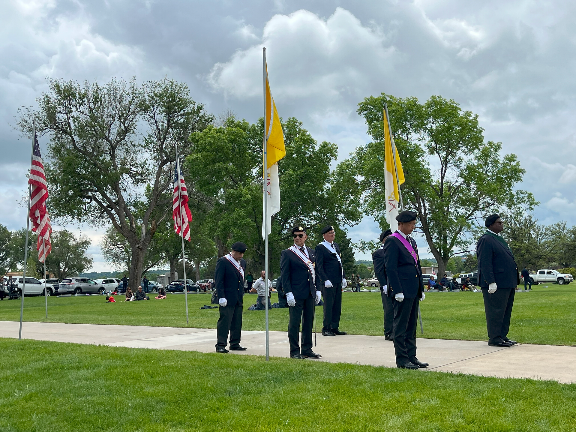 People in formal attire stand in line holding flags on a grassy lawn with trees. Day is cloudy, mood is ceremonial.