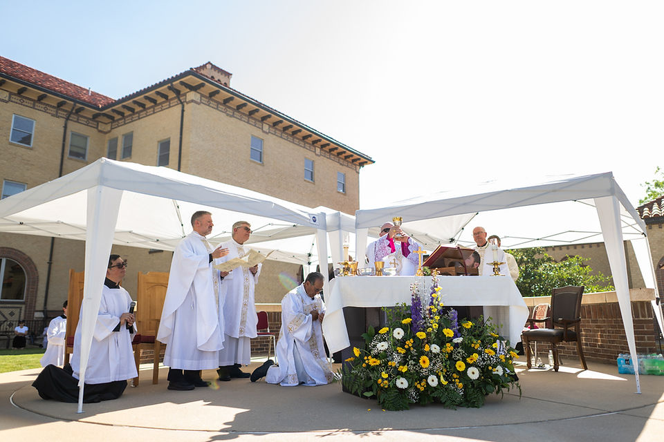 Outdoor Catholic Mass under white canopies, priests in white robes around the altar with flowers, chalice, and kneeling worshippers
