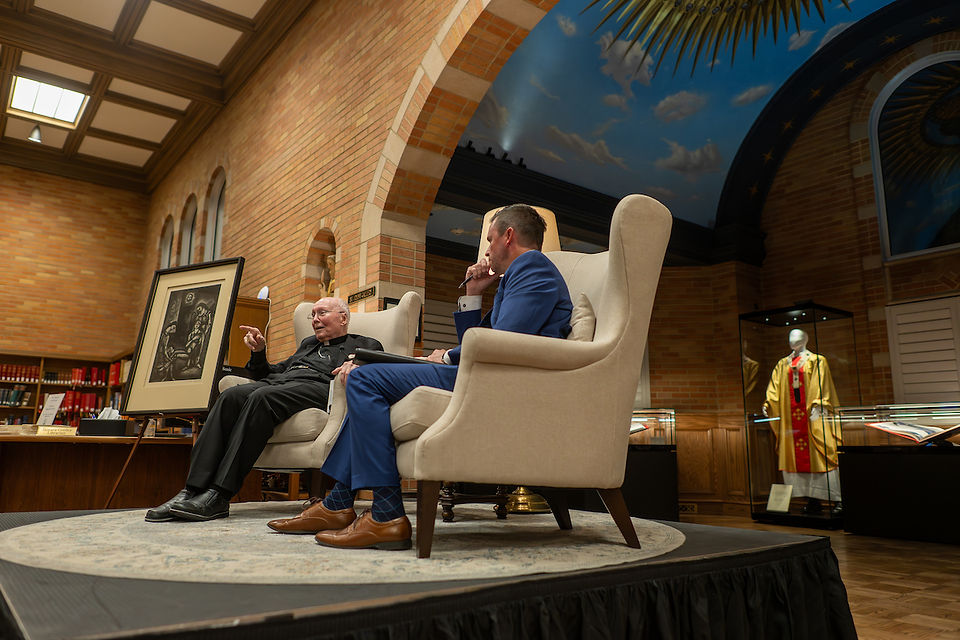 Two men sit conversing in a library with brick walls. A framed artwork and mannequin in robes are displayed. Ceiling has a sky mural.