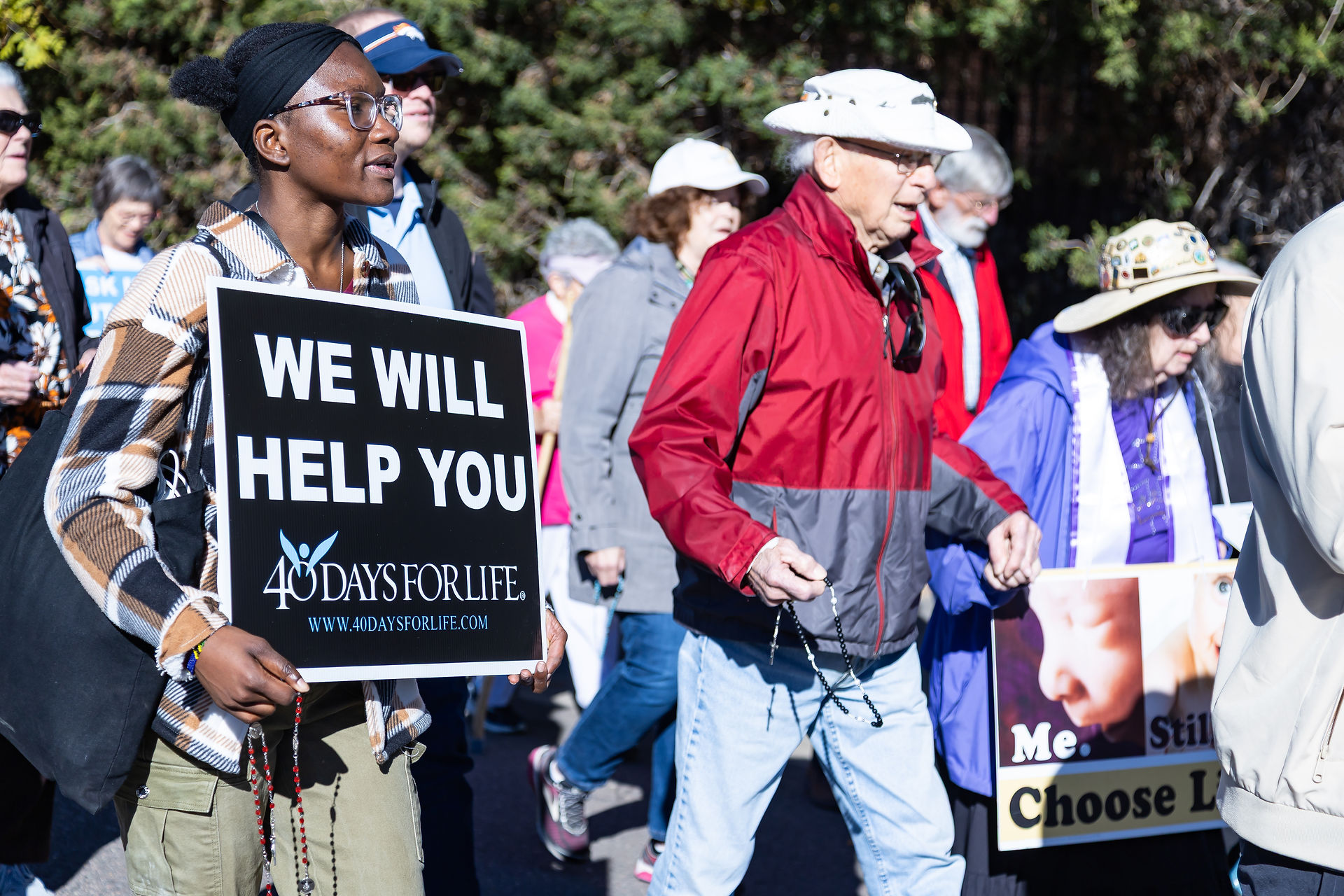 People marching outdoors holding signs and rosaries. A sign reads "We Will Help You, 40 Days for Life." The mood is determined.