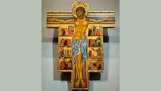 Crucifix with a golden Jesus, arms outstretched, surrounded by colorful religious scenes on a wooden cross, set against a gray wall.
